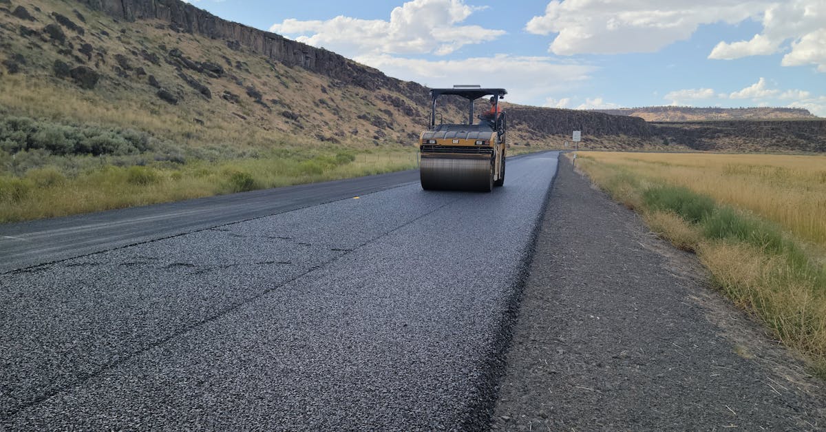A road roller paving a new asphalt road in a rural landscape under a blue sky.